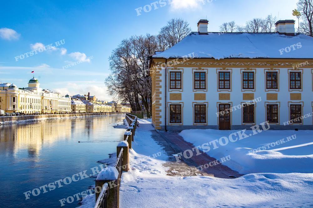 Фотография демонстрирует живописный зимний вид на историческое здание вдоль набережной реки Мойки в Санкт-Петербурге. Это летний дворец Петра Первого.